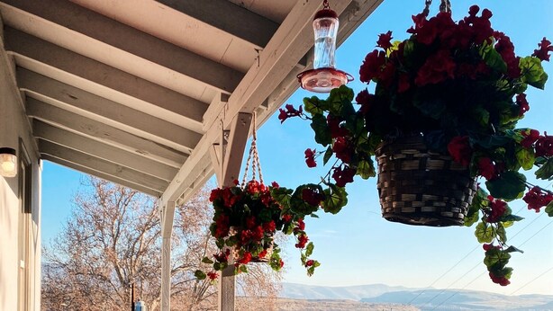 A wooden deck with a railing, a welcome mat, and hanging flower baskets. - Charming River View Apt | Walk to Shops & Cafes (The Dalles)