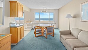 A compact living space with a kitchenette, a dining area with a round table and chairs, a sofa, and a large window offering a view of the ocean. - Rockaway Beach Resort (Rockaway Beach)