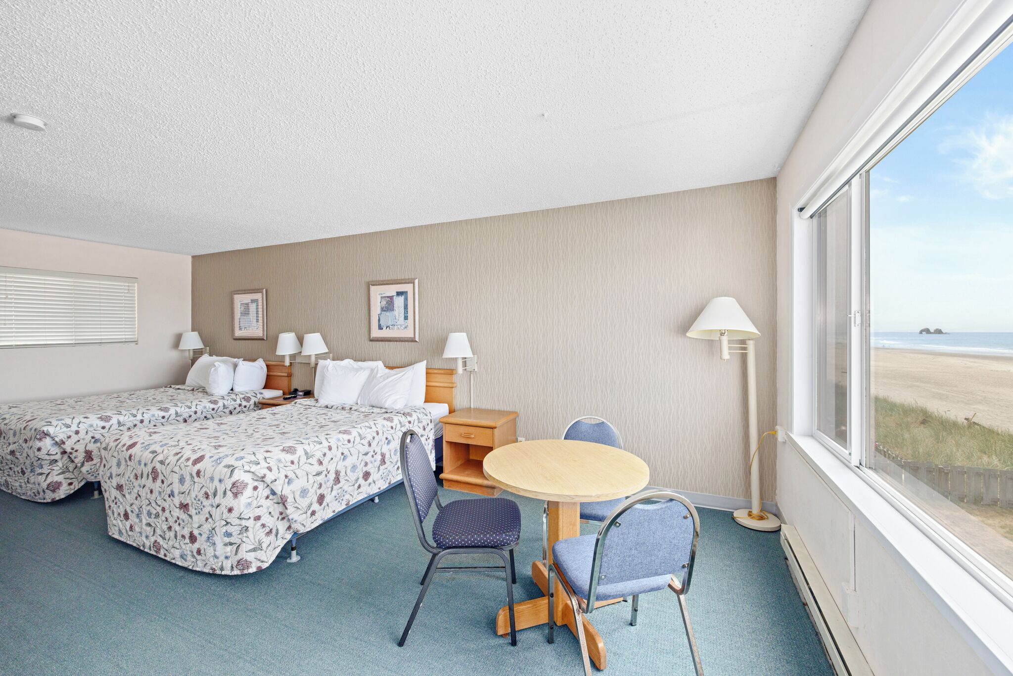 A hotel room with two beds, a small round table with chairs, a desk with a lamp, and a large window with a beach view.