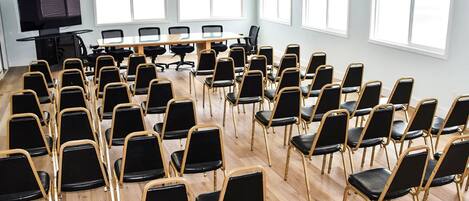 Un salón de conferencias con filas de sillas negras, una mesa larga y amplios ventanales.