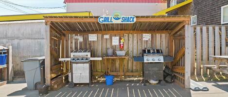 A small outdoor grill shack with two barbecue grills, a wooden table, and a covered seating area.