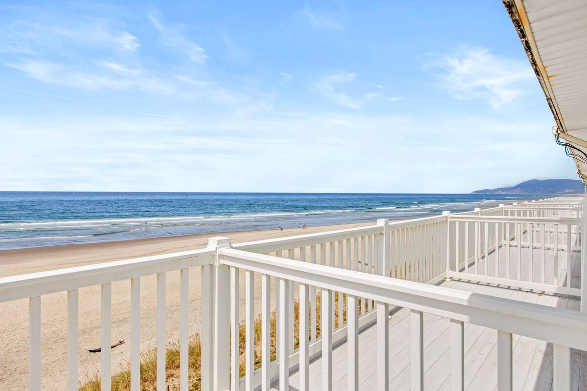 Vue sur la plage depuis une terrasse en bois dotée d’une balustrade, surplombant l’océan et la plage de sable.