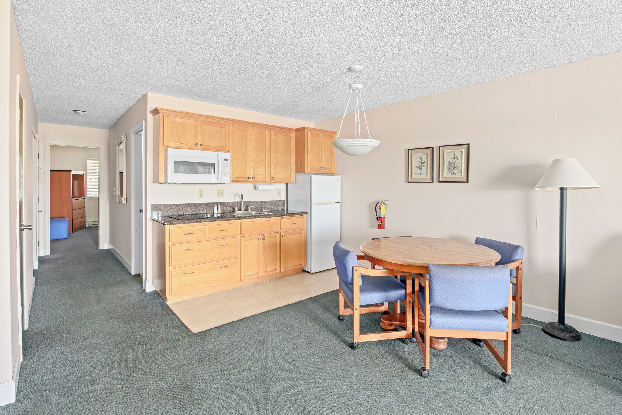 A compact kitchen with wooden cabinets, a microwave, and a refrigerator. There is a dining table with chairs and a floor lamp.