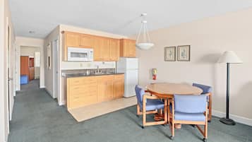 A compact kitchen with wooden cabinets, a microwave, and a refrigerator. There is a dining table with chairs and a floor lamp.