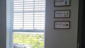 A window with horizontal blinds, a view of a street with cars, and three framed signs on the wall.
