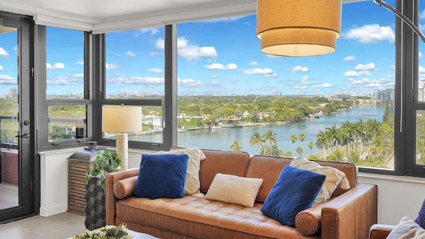Un salon moderne avec un canapé en cuir, une lampe et une vue sur une rivière et le paysage urbain, le tout visible à travers de grandes fenêtres.