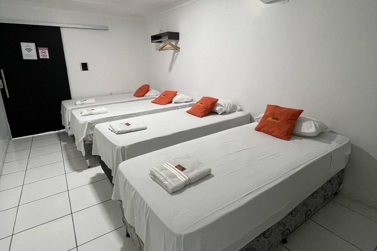 A hotel room with four single beds, each with white linens and orange pillows. There is a black door on the left, an air conditioning unit on the wall, and a small shelf above the beds.