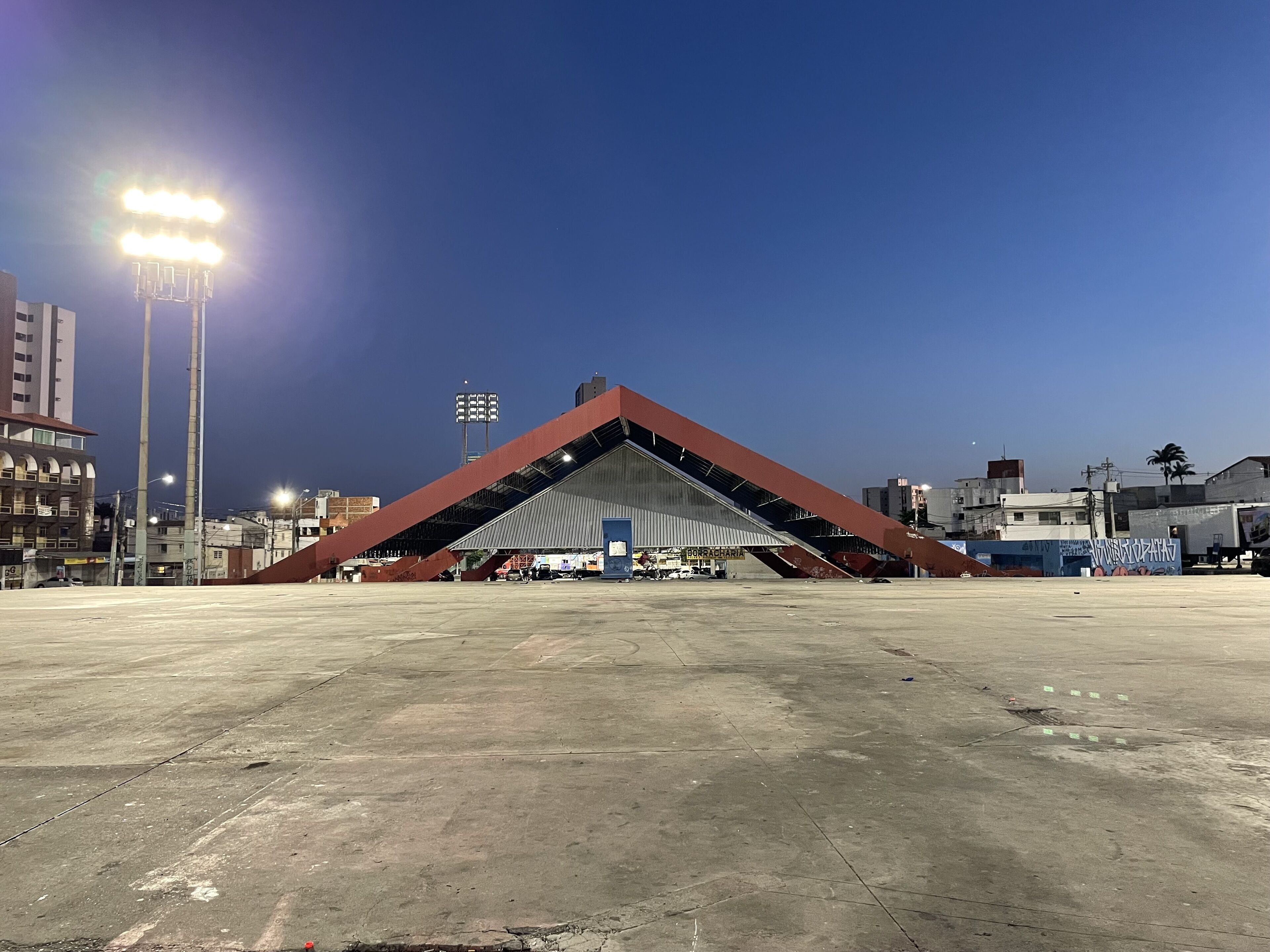 A large, open concrete area with a prominent red structure in the center, possibly a stadium or sports facility, surrounded by buildings and streetlights.