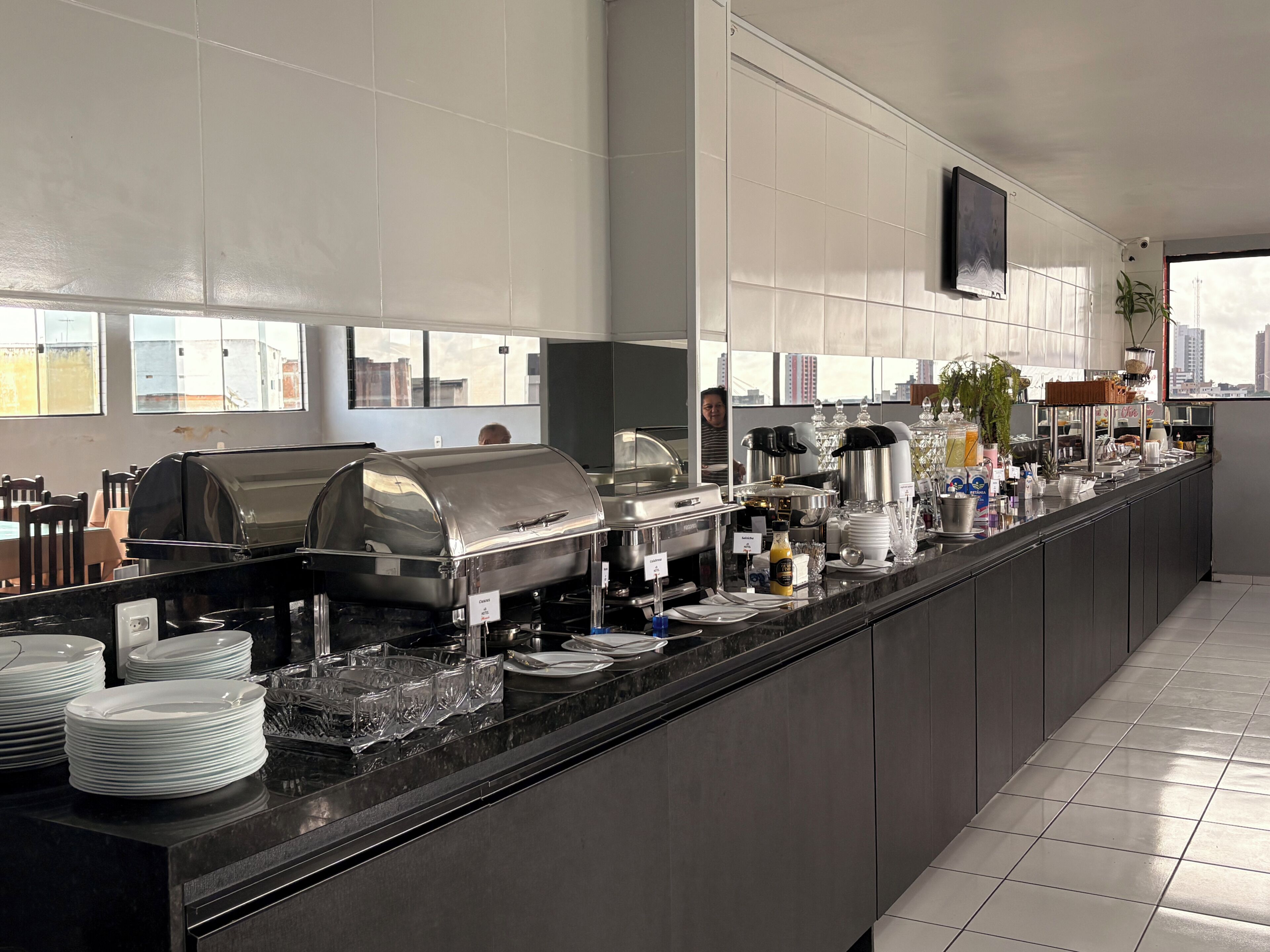 Et moderne hotelkøkken med buffet, inklusive selvbetjeningsstation, kaffemaskine og et stort vindue med udsigt over byen.