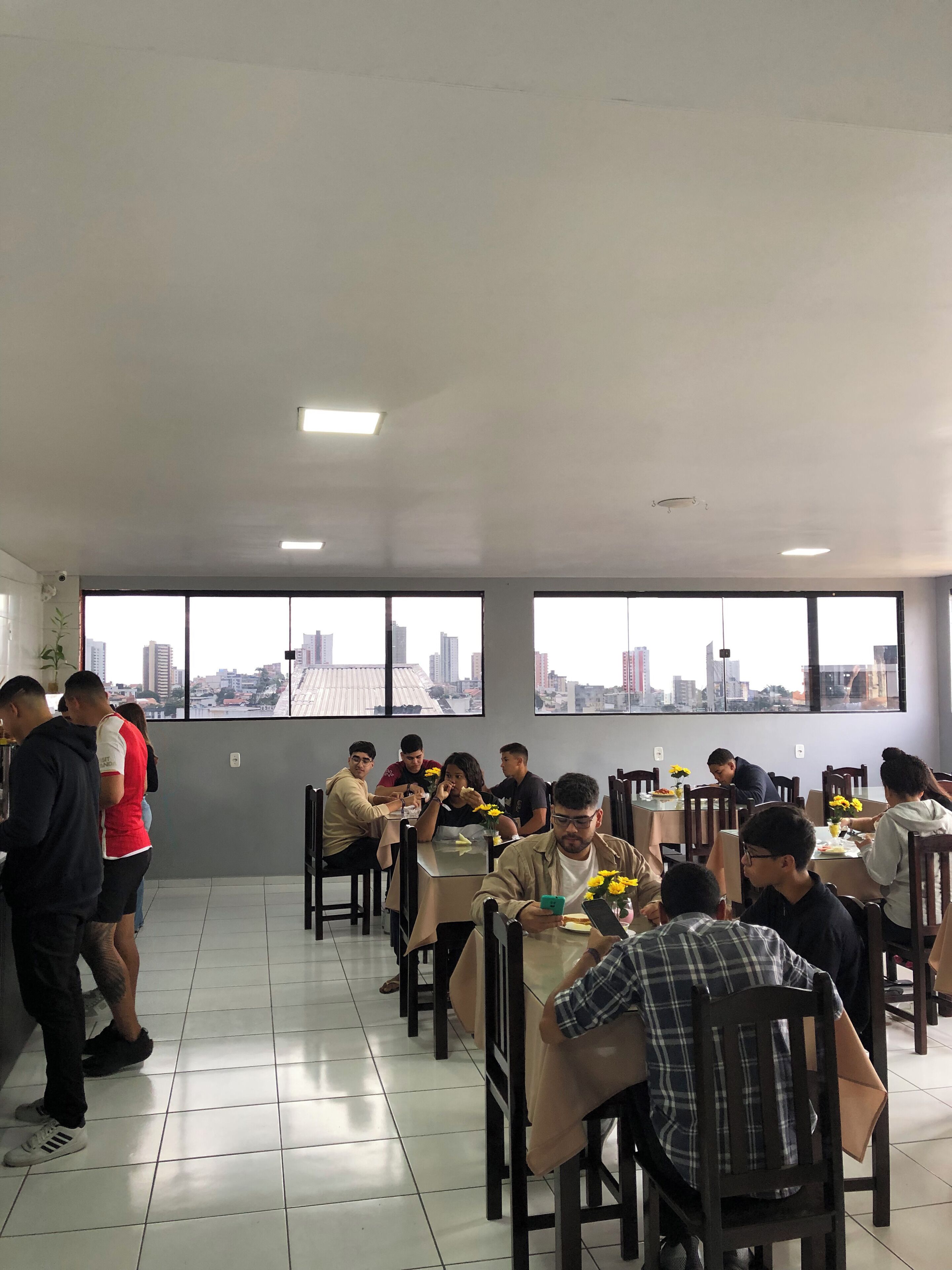 A dining area with tables and chairs, large windows with a city view, and a tiled floor.