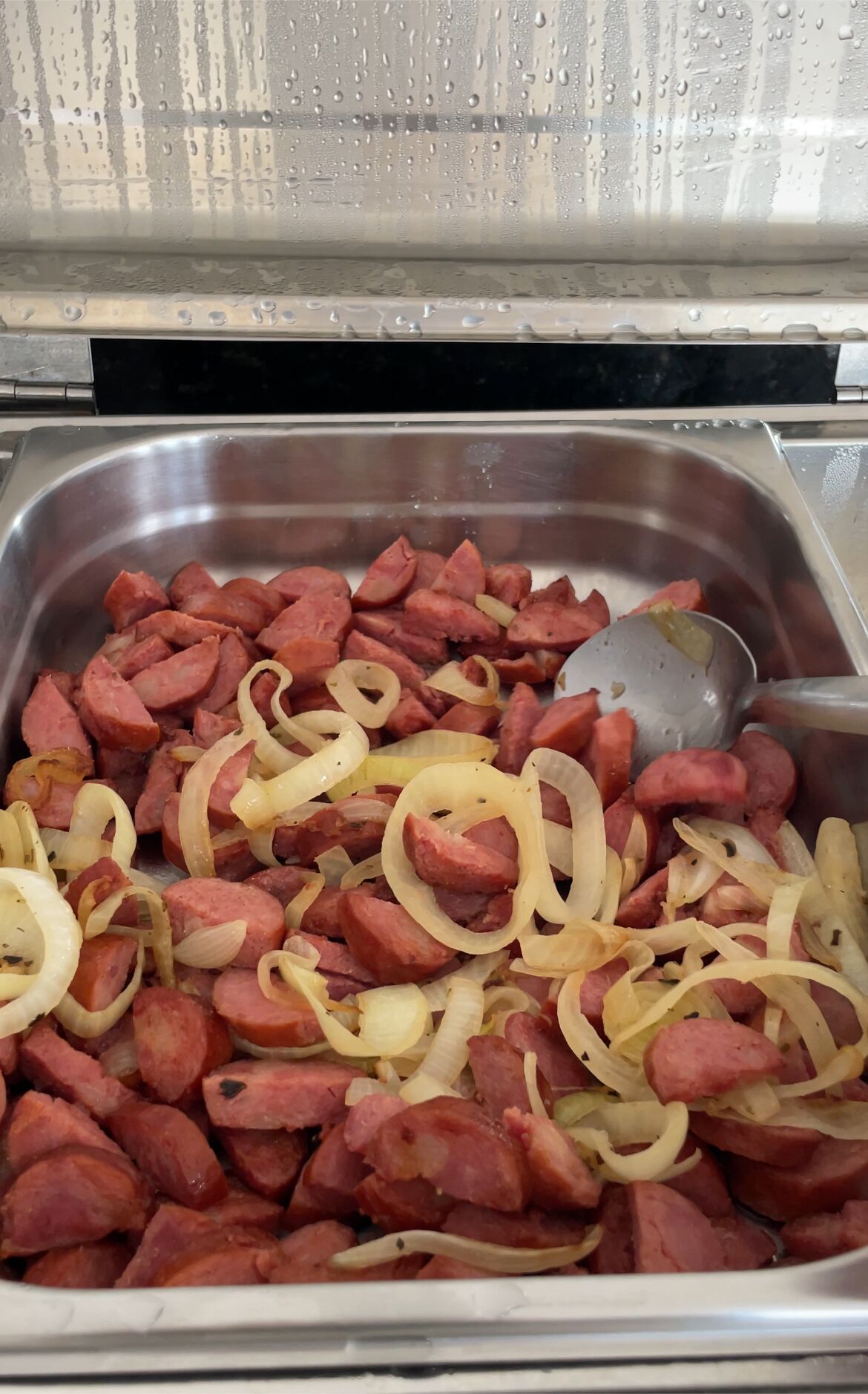 A stainless steel buffet dish containing sliced sausages and onions.