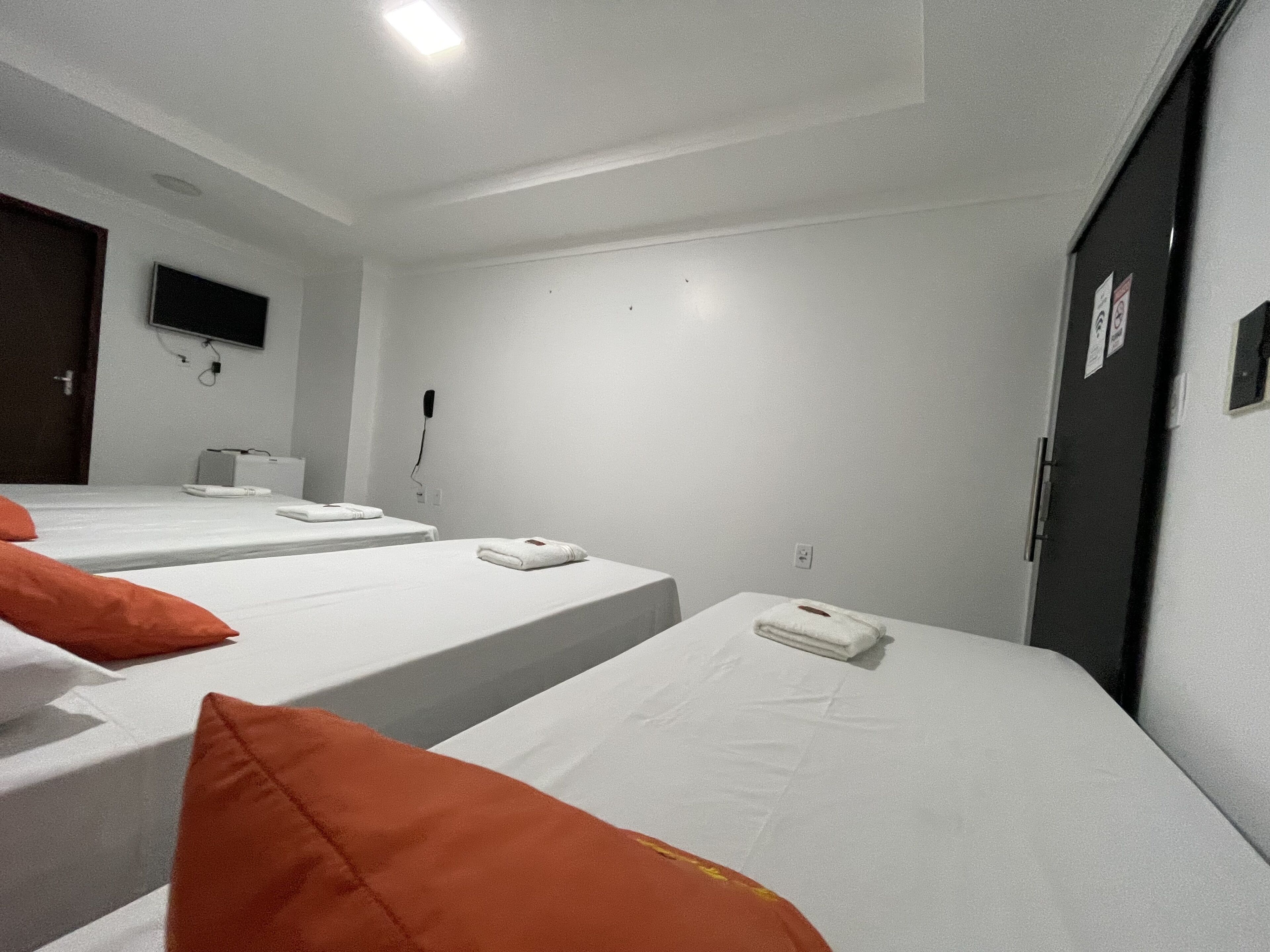 A hotel room with three beds, each with white bedding and orange pillows. There is a flat-screen TV mounted on the wall, a power outlet, and a door with a sign.