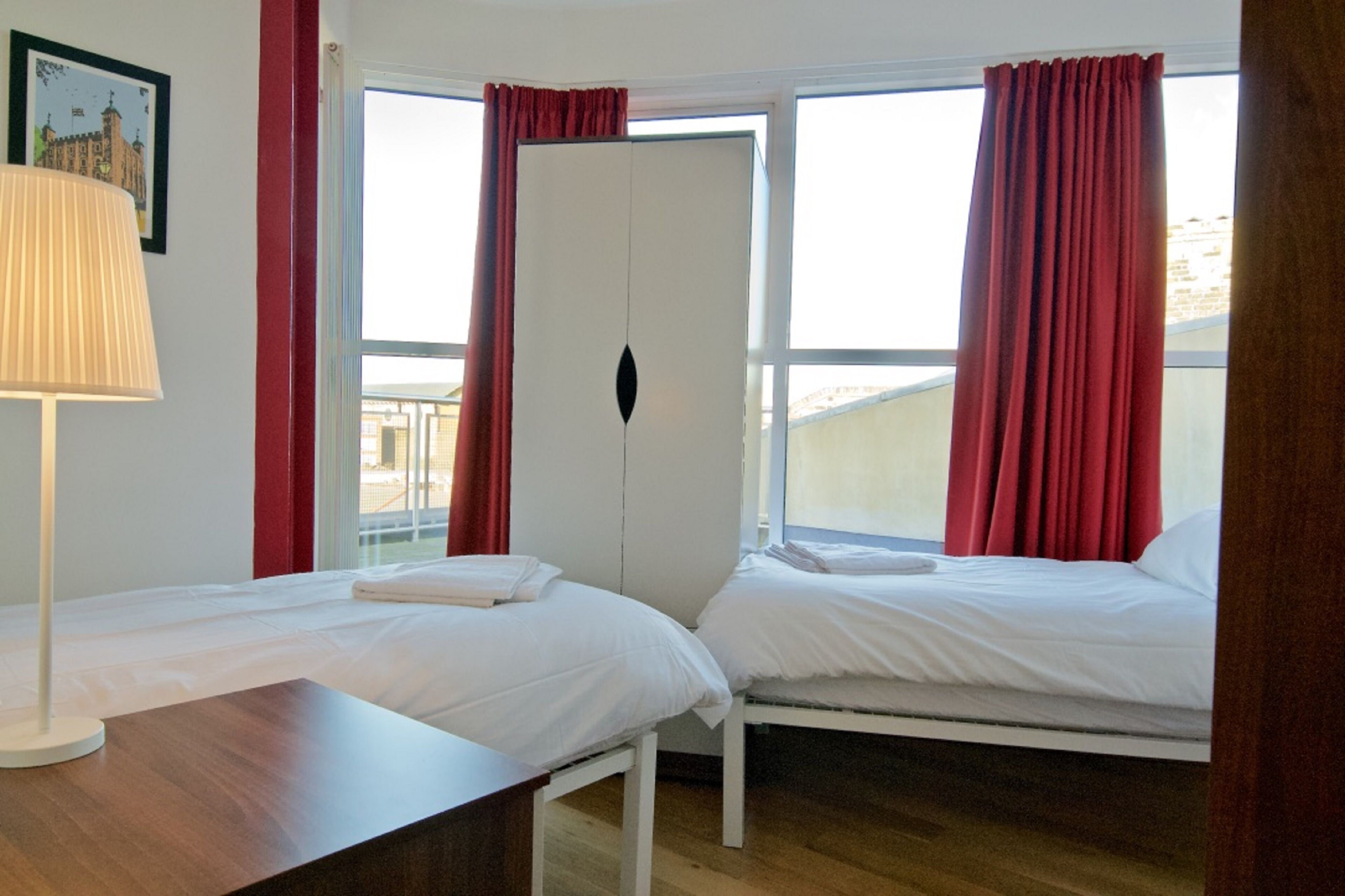 A hotel room with two beds, a wardrobe, a window with red curtains, and a lamp.