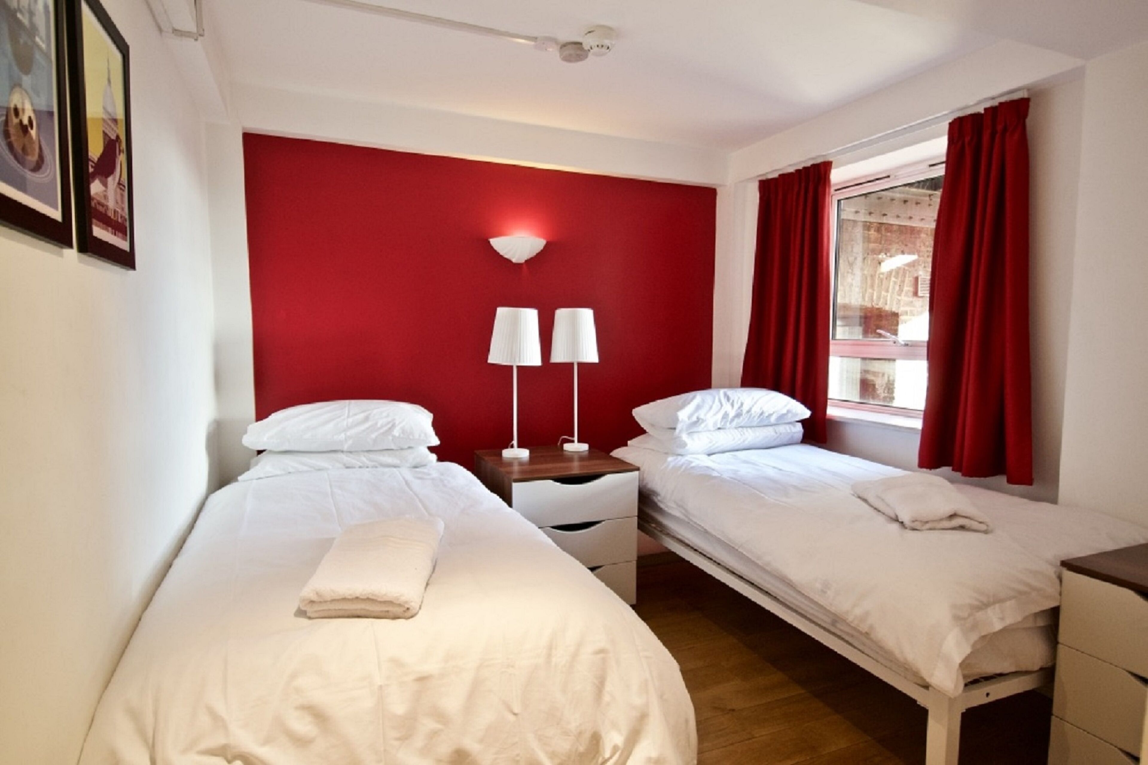 A hotel room with two single beds, a red accent wall, white bedding, two bedside lamps, and a window with red curtains.