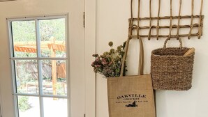 A room with a white door, a window, a wall-mounted wooden rack with bags, a woven basket, and a copper kettle on a table. - Chic wine country cottage and garden (Healdsburg)