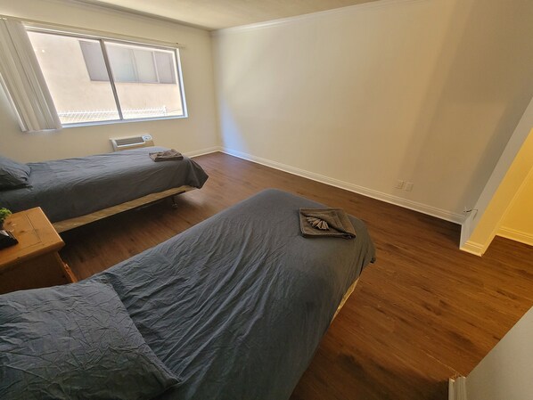 Two single beds, a bench, and a table with a potted plant, set against a light-colored backdrop with a balcony view.