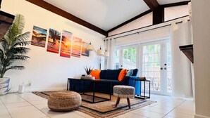 A spacious living room with a dark blue sofa, orange cushions, a patterned ottoman, and a large potted plant.