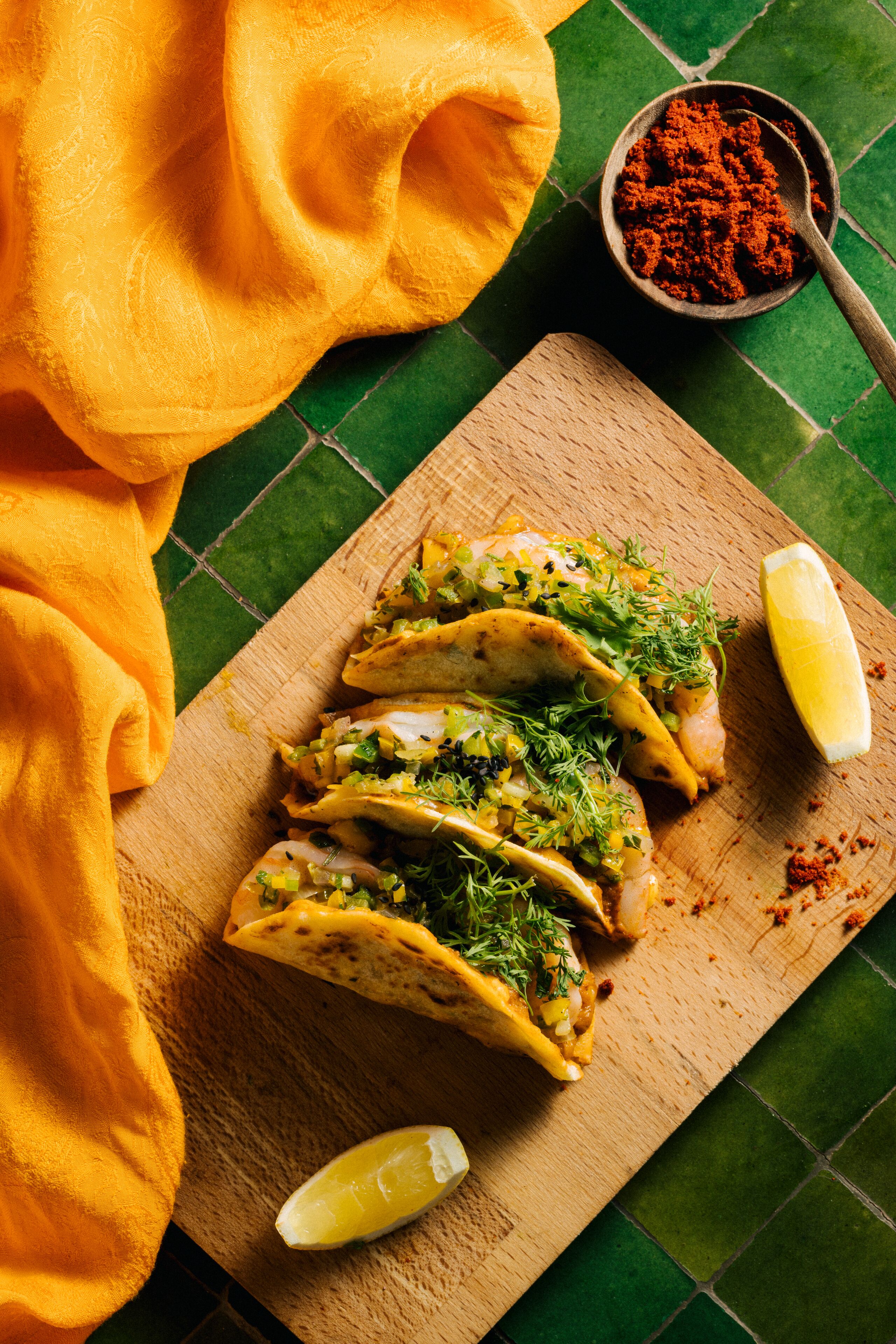Tres tacos sobre una tabla de madera con un gajo de limón y un tazón de condimento rojo.