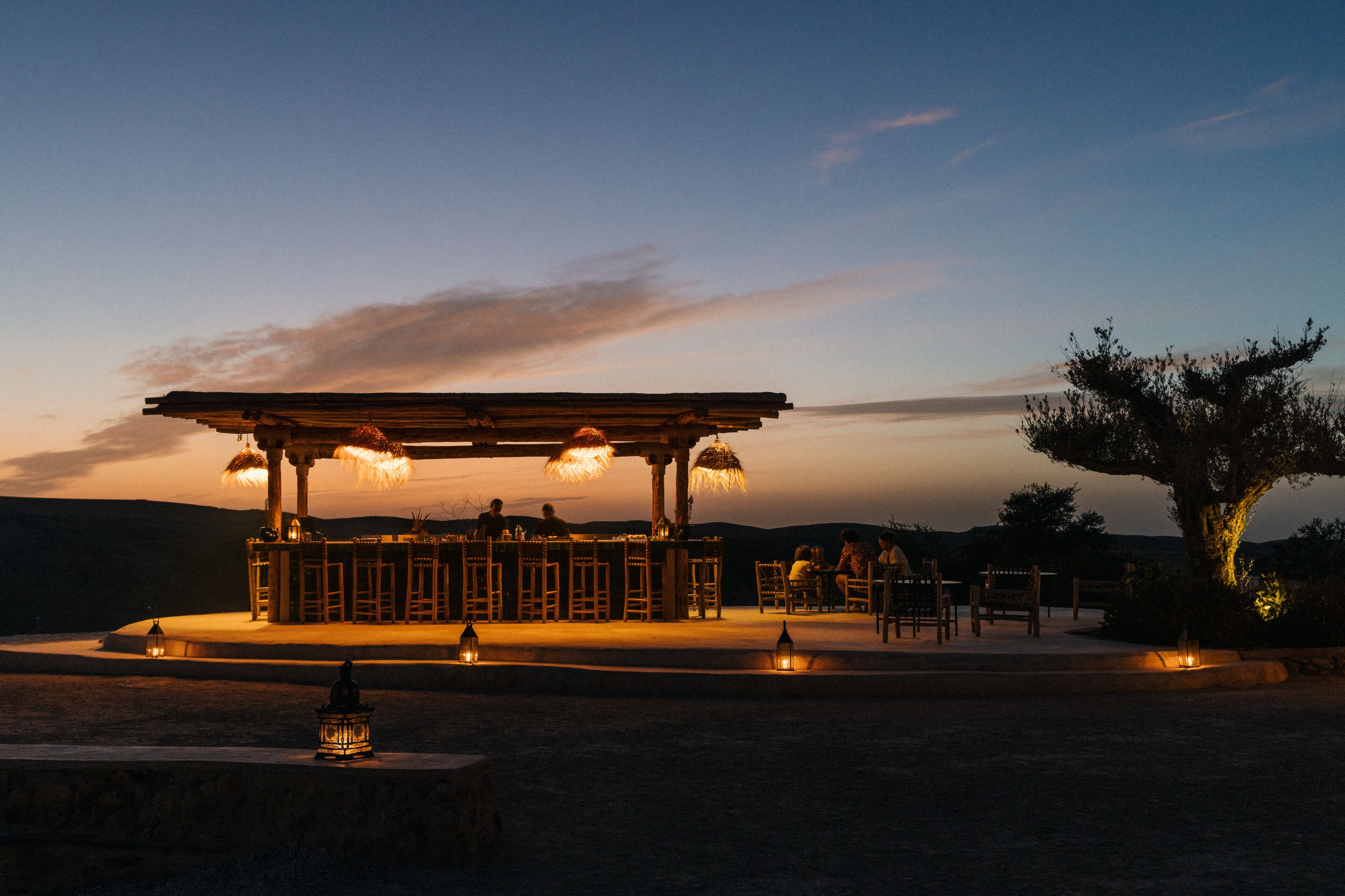 An outdoor seating area with a wooden pergola, tables, and chairs, surrounded by a landscaped garden.