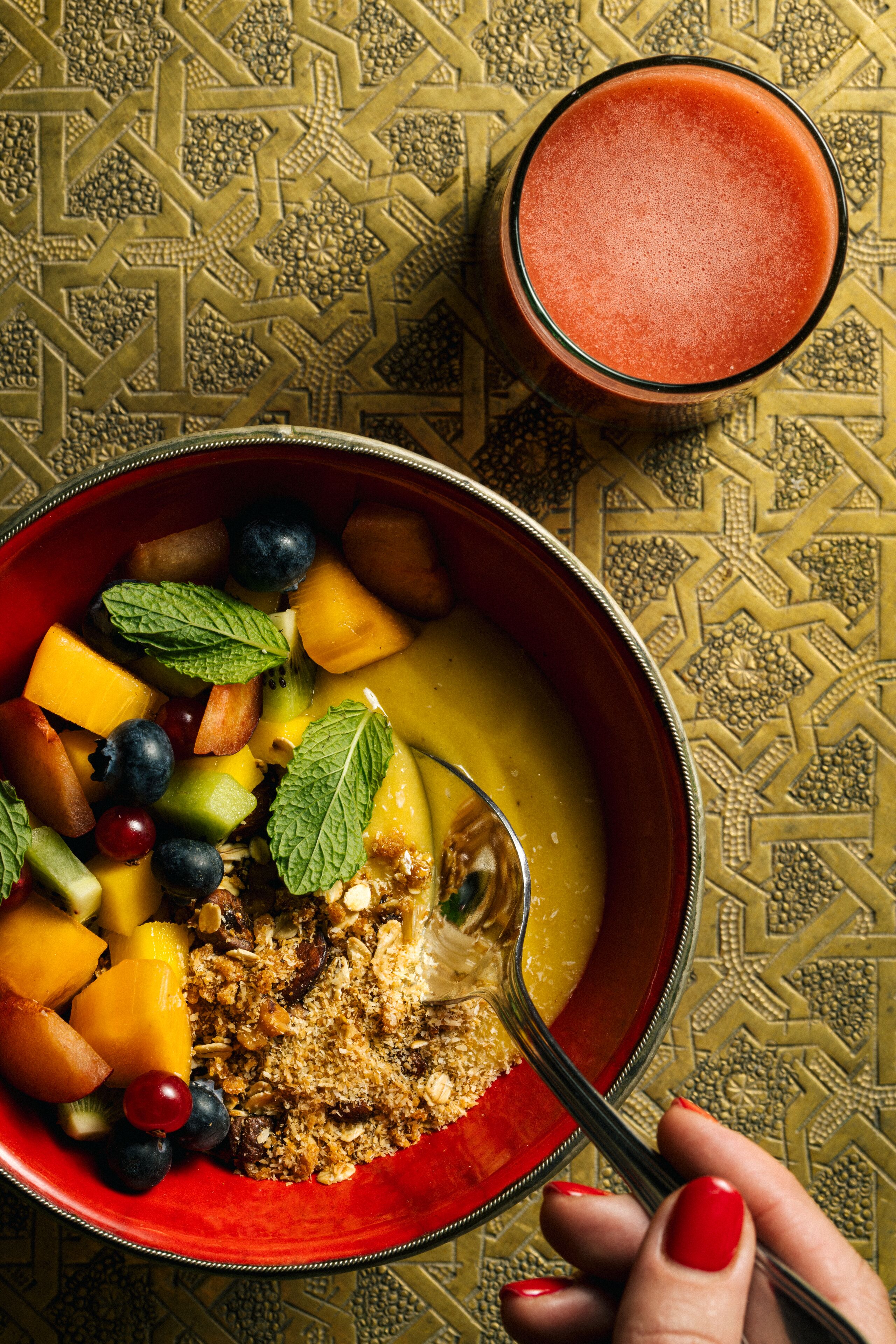 A bowl of fruit salad with a spoon, a glass of smoothie, and a patterned tablecloth.