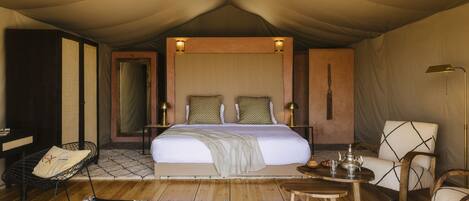 A tented room with a bed, bedside tables, a chair, a small table, and a lamp.