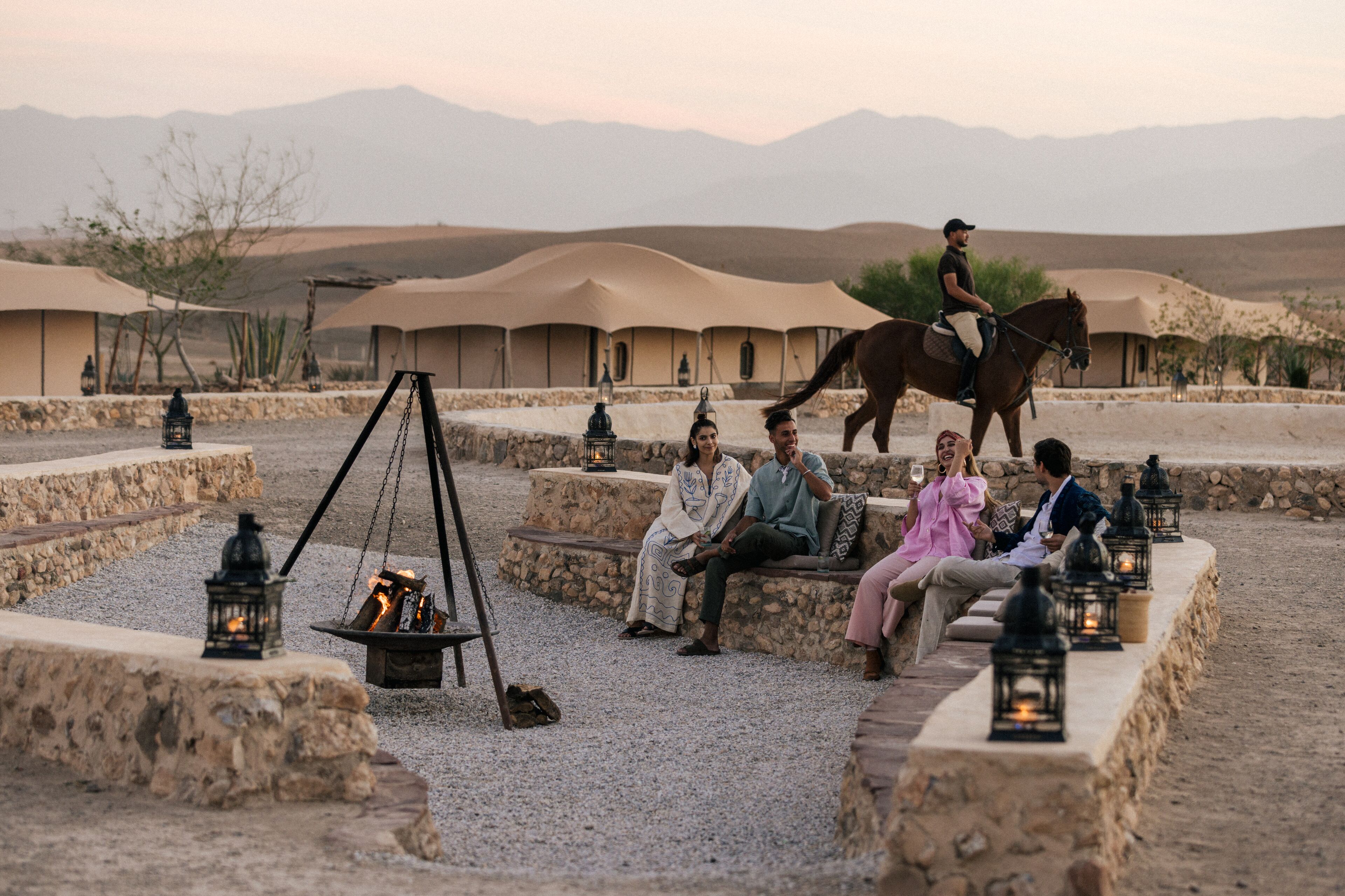 A desert setting with a group of people sitting on stone benches around a fire pit, with a horse and rider in the background.