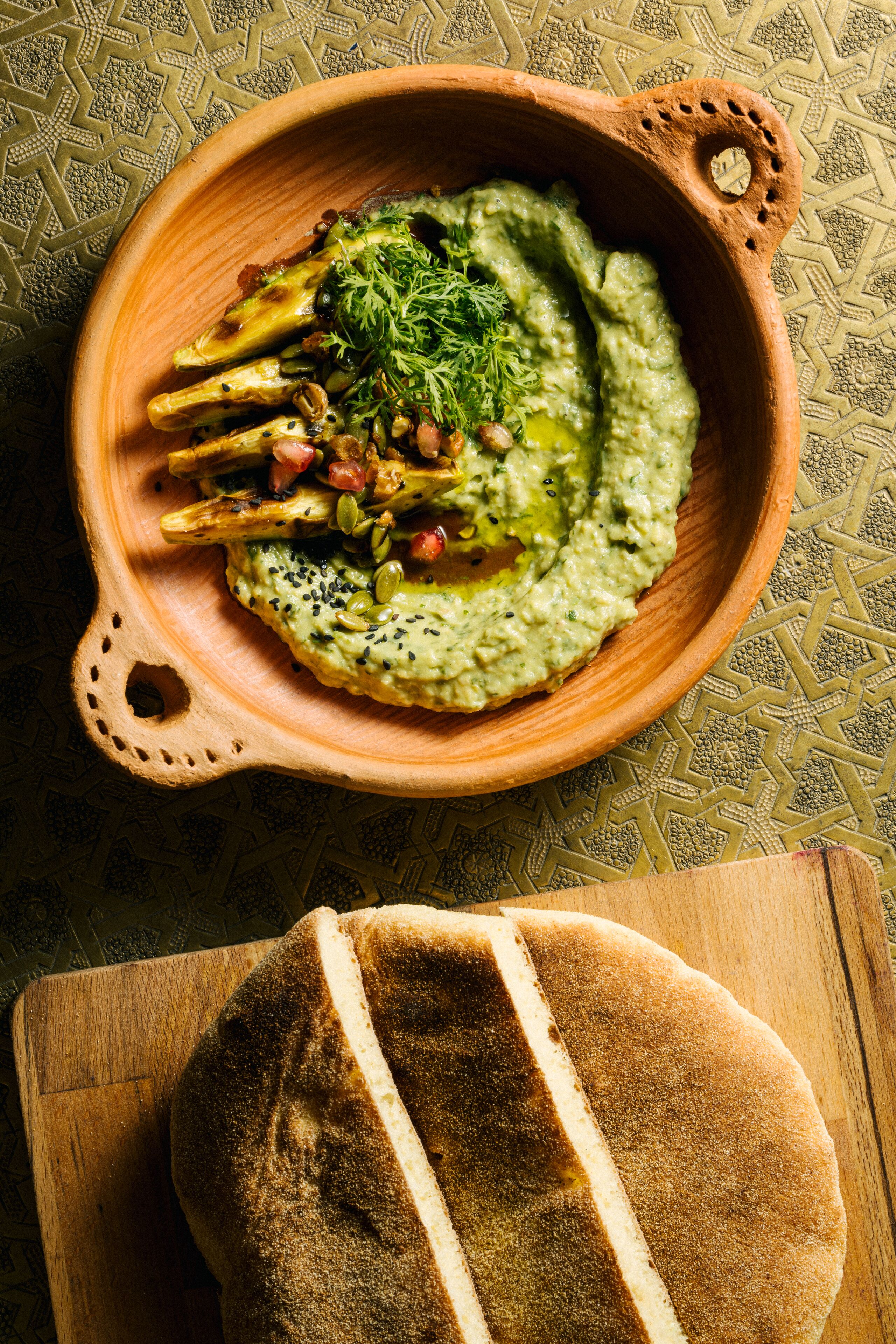 A wooden bowl with a green sauce and vegetables on a patterned tablecloth.