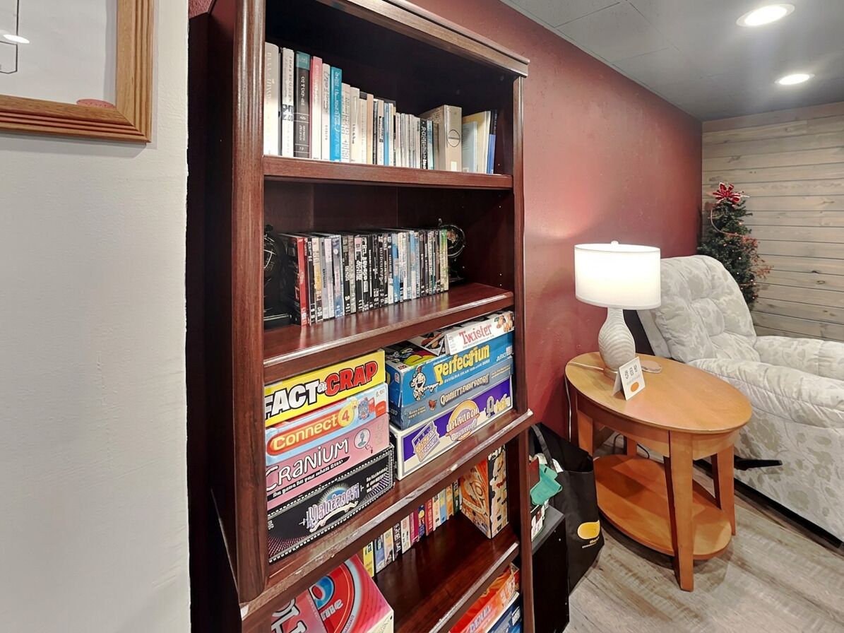 Ein Zimmer mit Bücherregal, das mit Büchern und Gesellschaftsspielen gefüllt ist, einem kleinen runden Tisch mit einer Lampe und einem weißen Sessel.
