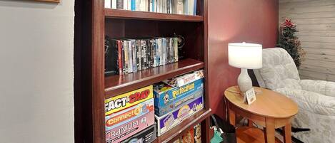 Ein Zimmer mit Bücherregal, das mit Büchern und Gesellschaftsspielen gefüllt ist, einem kleinen runden Tisch mit einer Lampe und einem weißen Sessel.