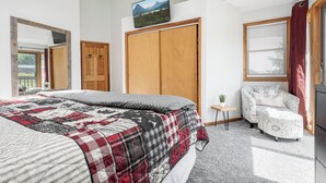 A bedroom with a large bed, a wooden wardrobe, a television on the wall, a chair, and a small table with a plant.