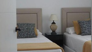 A hotel room with two beds, each with a headboard, a bedside table with a lamp, and a blanket on one bed. - HOLIDAY BEACH VACATIONS (El Puerto de Santa María)