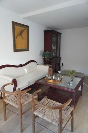 Un salon avec un canapé blanc, deux chaises en bois à assise tressée, une table basse en verre agrémentée d’objets décoratifs, un meuble en bois foncé et un tableau encadré accroché au mur.