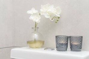 Bathroom features a clear glass vase with white flowers and two glass candle holders on a minimalist surface.