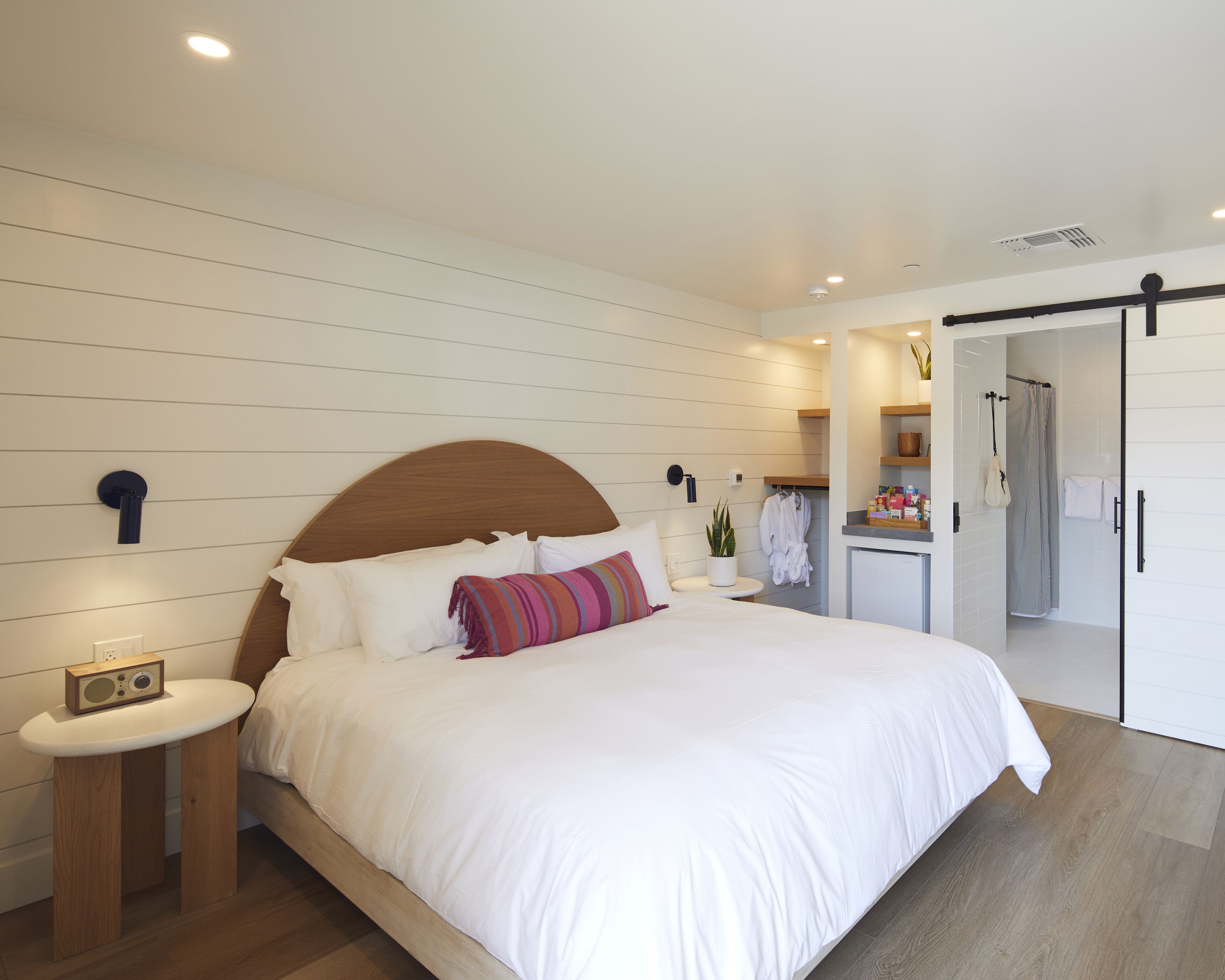 Un dormitorio moderno con cabecera de madera, una mesita de noche con radio, una cama grande con ropa de cama blanca y un vestidor con ducha.