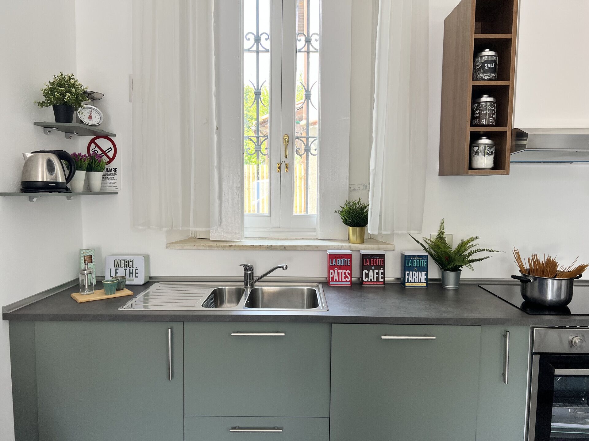 Una cucina con lavello, mobili, una finestra con tende e uno scaffale con utensili da cucina.