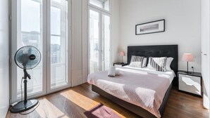 A bedroom with a large bed, a standing fan, and a city view through the windows. - GuestReady - Bright Apartment near Bolhão (Porto)