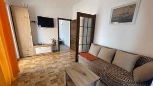 Sala de estar con un sofá estampado, una mesa de centro de madera, un televisor montado en la pared y una zona de cocina con armarios y una ventana.