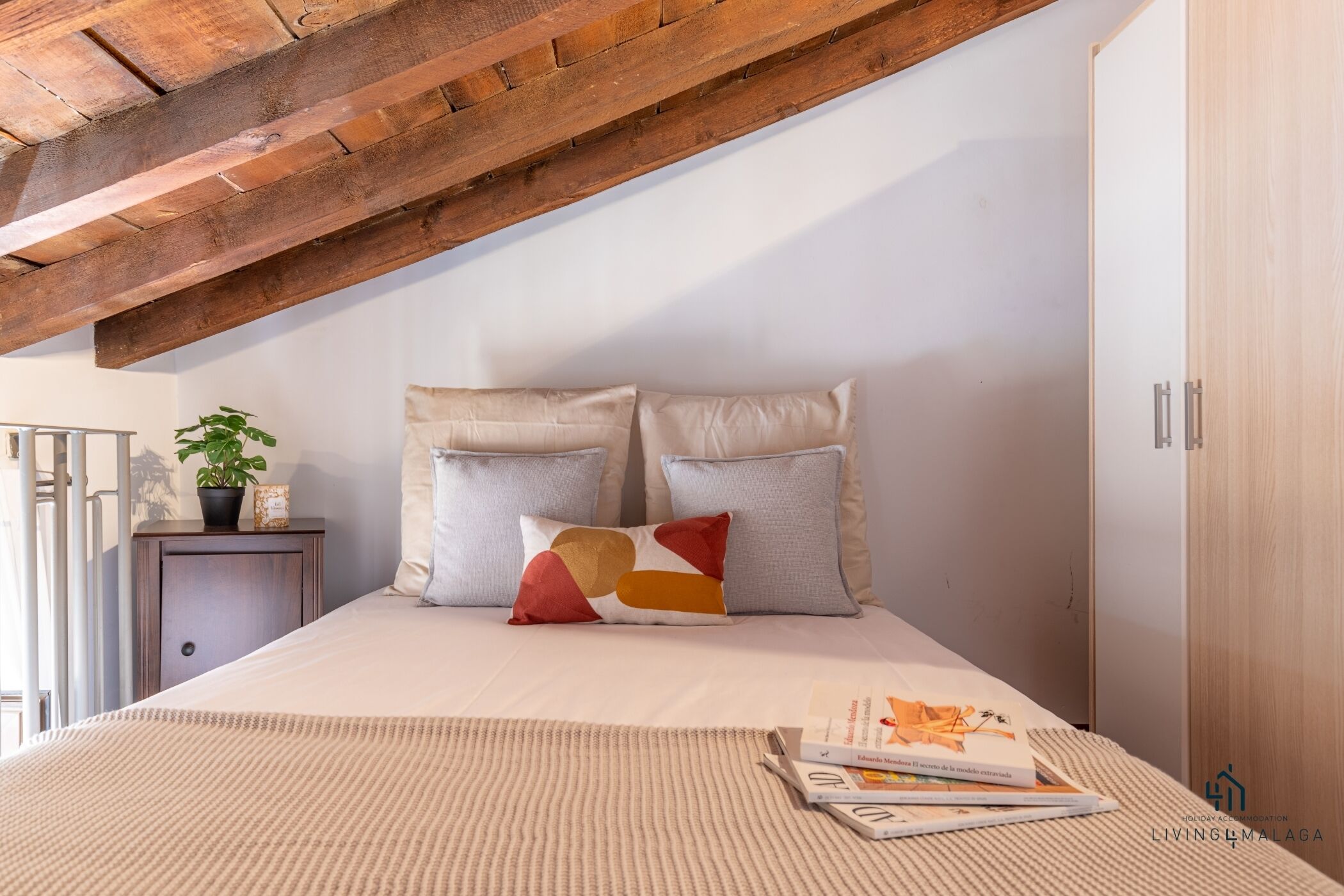Ein Schlafzimmer mit schräg stehender Decke, einem Bett mit beigefarbener Bettwäsche, zwei Kissen, einem kleinen Nachttisch mit Pflanze und einer Zeitschrift.