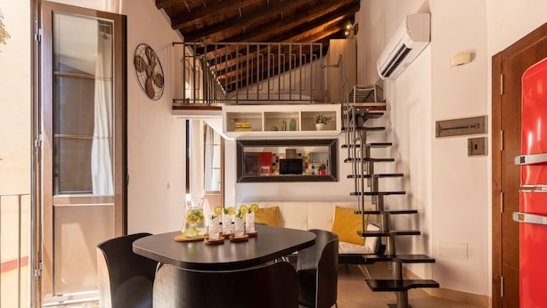 A modern kitchen with a black countertop, a dining area with black chairs, a staircase, and a red door.