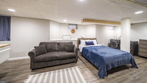 Un dormitorio con un sofá gris, una cama con colcha azul, una lavadora y suelo de madera.
