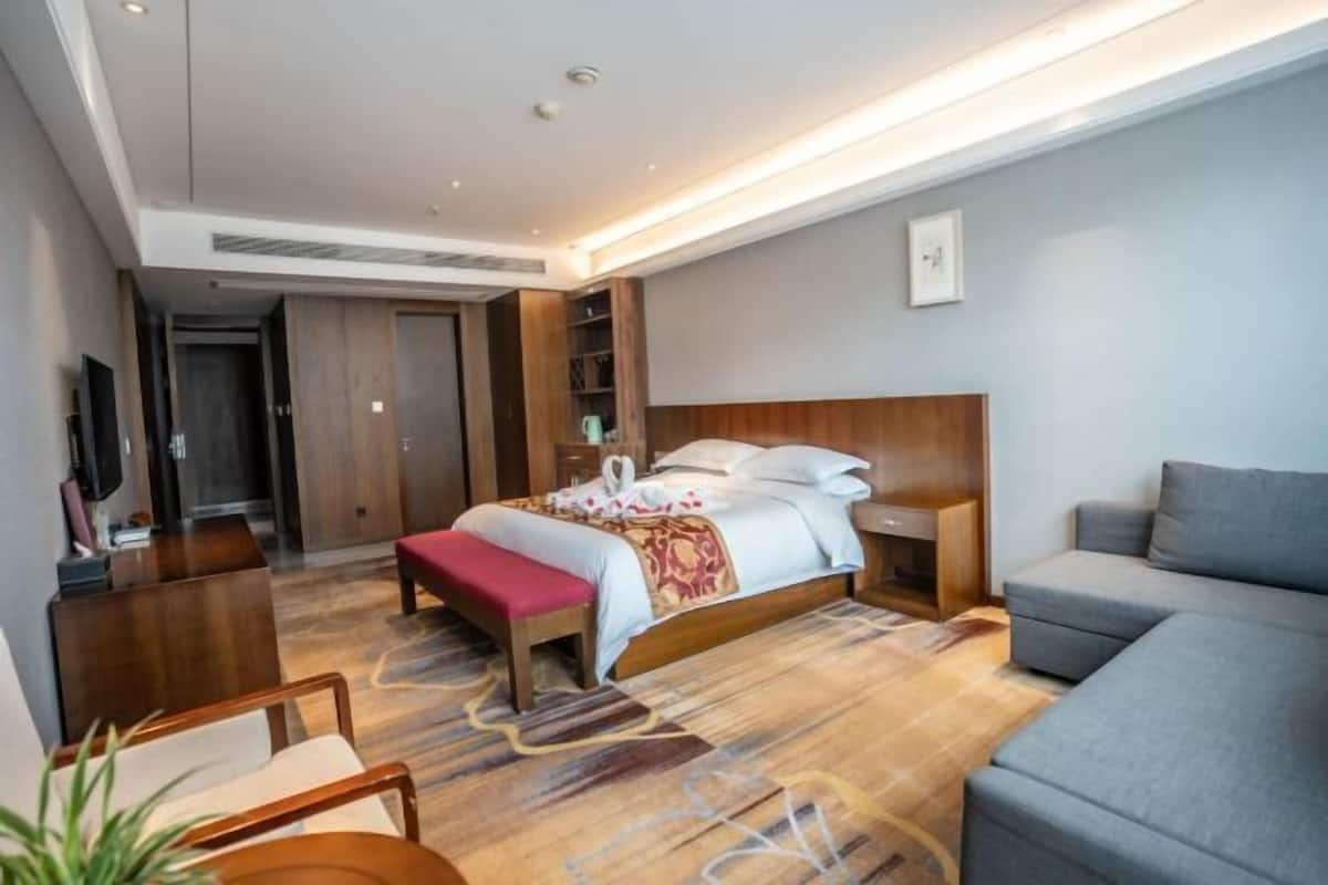 A modern hotel room with a large bed, a grey sofa, a wooden bench, and a TV on a wooden stand.