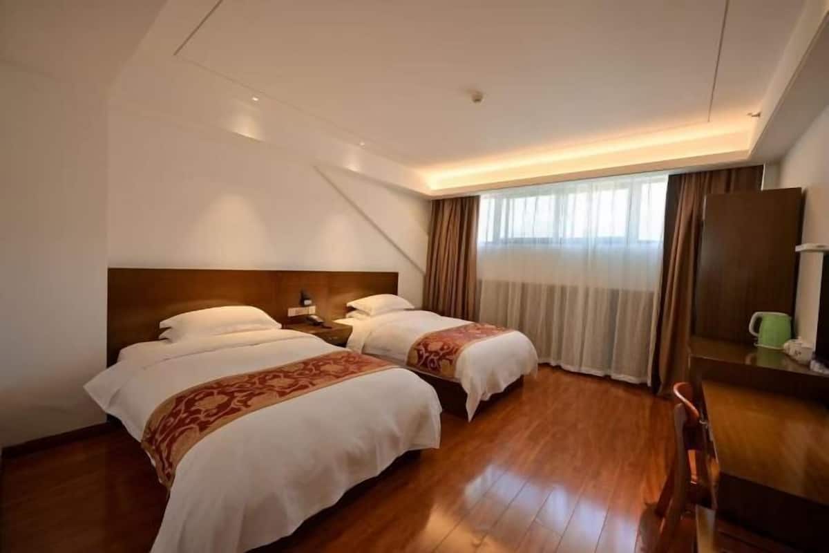 A hotel room with two beds, a wooden floor, a large window with curtains, and a desk with a lamp.