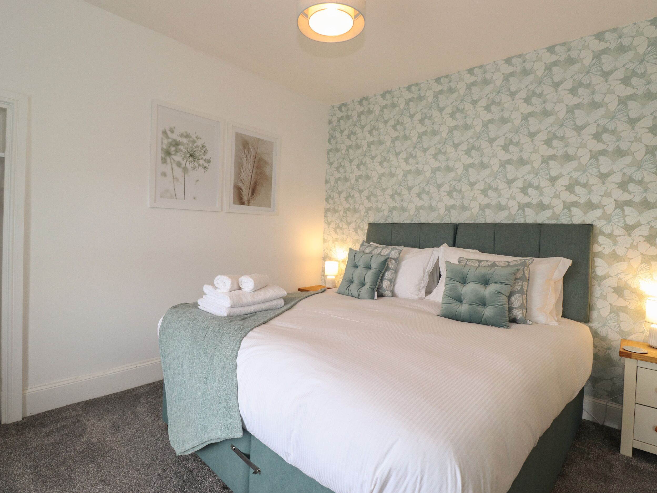 Un lit bien fait, avec une tête de lit verte, une literie blanche et des coussins décoratifs. On y trouve deux tables de chevet avec des lampes, et une pile de serviettes pliées posée sur le lit.