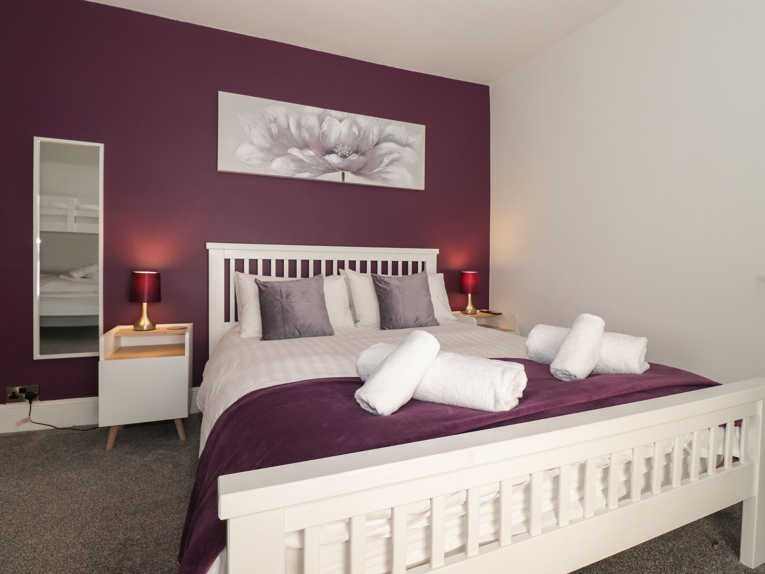 Un lit bien fait, avec une literie blanche et un plaid violet, deux lampes de chevet, une tête de lit blanche et une grande œuvre d’art florale accrochée au mur.