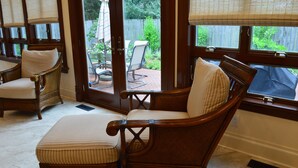 A room with a wooden chair and ottoman, a window with blinds, and a view of a backyard with patio furniture. - Welcome! Cozy large house to make memories of laughter and joy by the Big Blue! (South Haven)