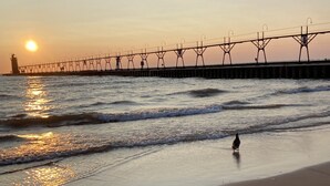 A beach with a pier extending into the water, a person walking along the shore, and the sun setting in the background. - Welcome! Cozy large house to make memories of laughter and joy by the Big Blue! (South Haven)