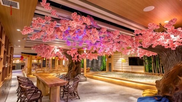 En restaurant med træpaneler, et langt bord med stole, og et dekorativt træ med lyserøde blomster.