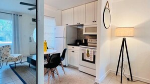 A compact kitchen with white cabinets, a microwave, and a stove. There is a dining area with a table and chairs, and a floor lamp. - Cozy 2 BR Condo - 7 Min from Fort Lauderdale Beach (Fort Lauderdale)