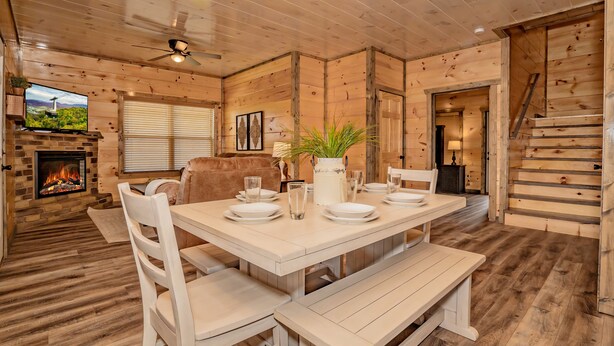 Ein gemütliches Blockhaus-Interieur mit einem Esstisch, gedeckt für vier Personen, einem Kamin, einem Fernseher und einer Treppe.