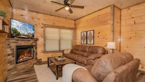 Ein gemütliches Blockhaus-Interieur mit Kamin aus Stein, Ledersofa und einem Fernseher, der ein Riesenrad zeigt.
